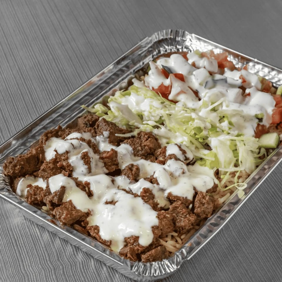 Mediterranean rice bowl with meat, lettuce, tomatoes, and white sauce in a foil container.